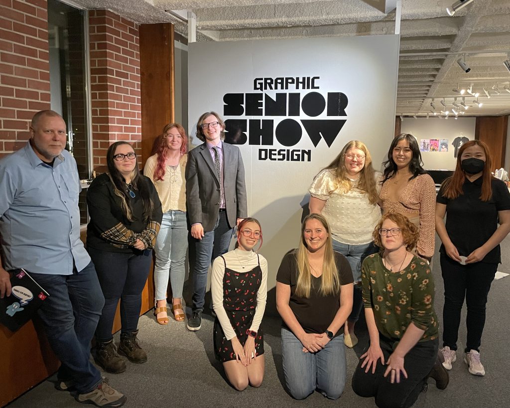 A photo of all the graphic design seniors together with professor Decker in front of a Graphic Design Senior Show sign.